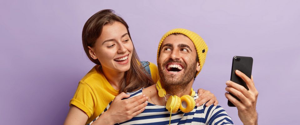Funny young couple take selfie on smartphone, enjoy piggyback ride, have happy expressions, lovely woman hugs boyfriend from back, isolated over violet background. People, fun, leisure time concept